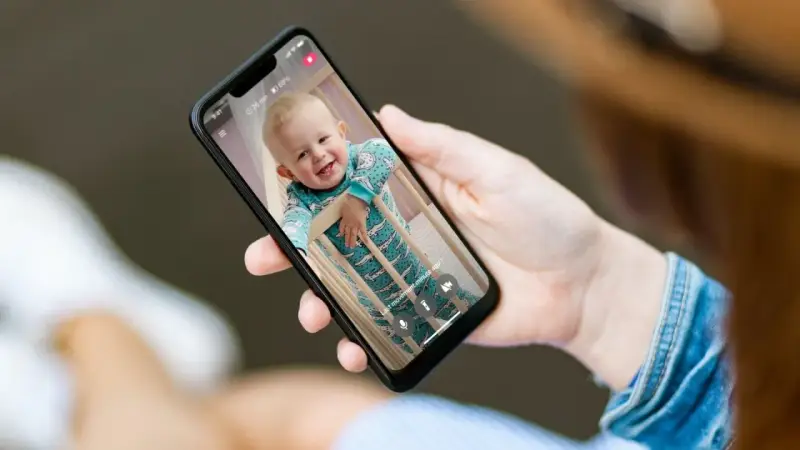 Ein lächelndes Baby im Strampler schaut aus dem Gitterbett. Eine Hand hält das Handy als Babyphone, um das Kind im Blick zu behalten.