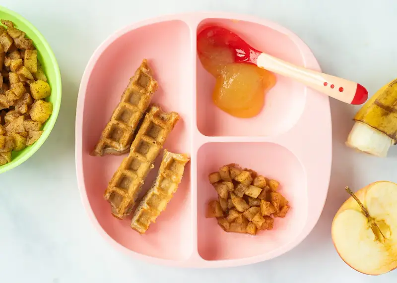 Baby-freundliche Apfelwaffeln mit Apfelmus und Zimt-Apfelstückchen auf einem rosa Teller. Ein halber Apfel und eine Banane liegen daneben.